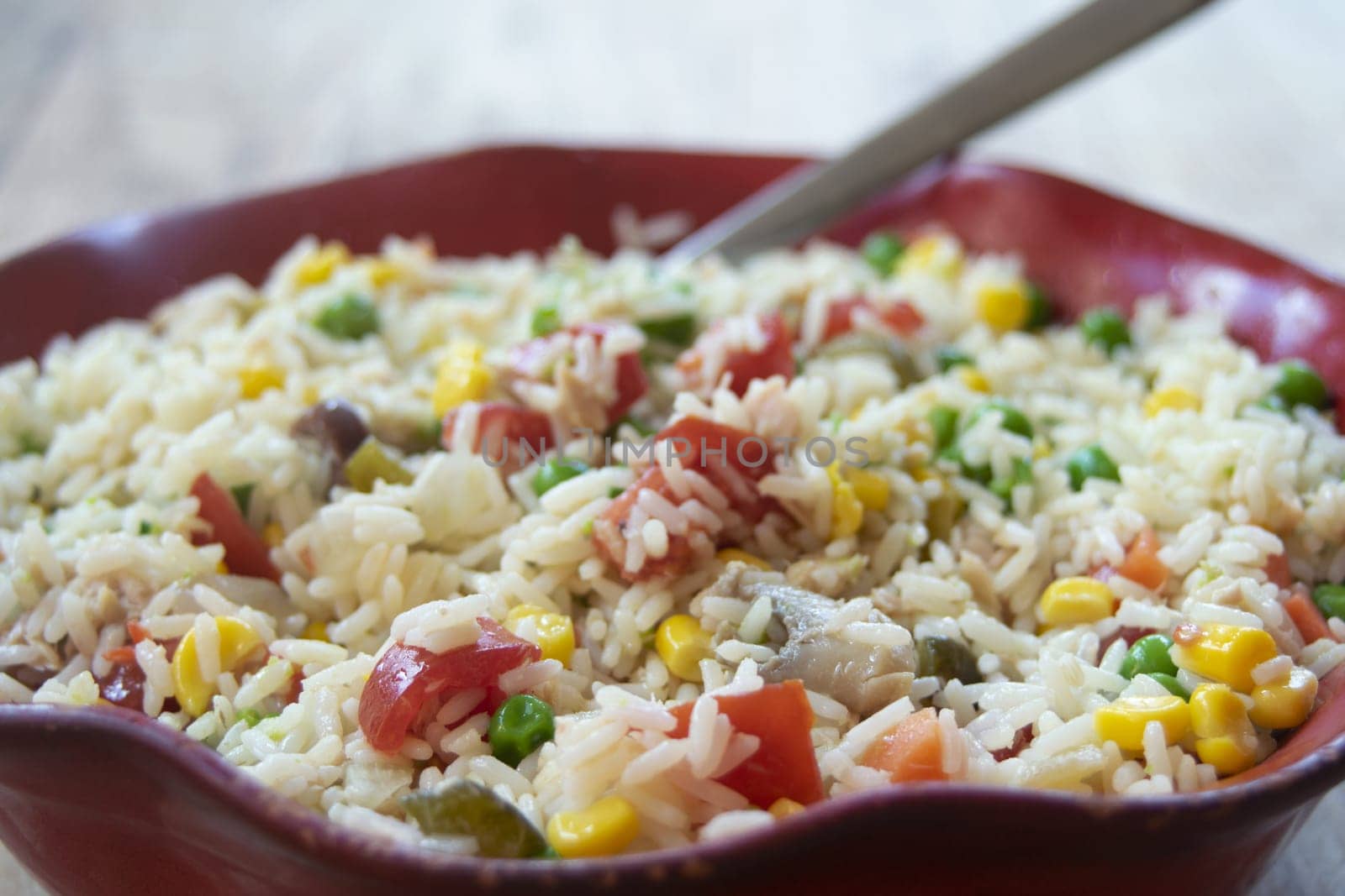 Close-up of a vibrant rice salad in a red bowl, featuring rice mixed with colorful vegetables like tomatoes, corn, peas, and possibly others. A spoon rests inside the bowl.

