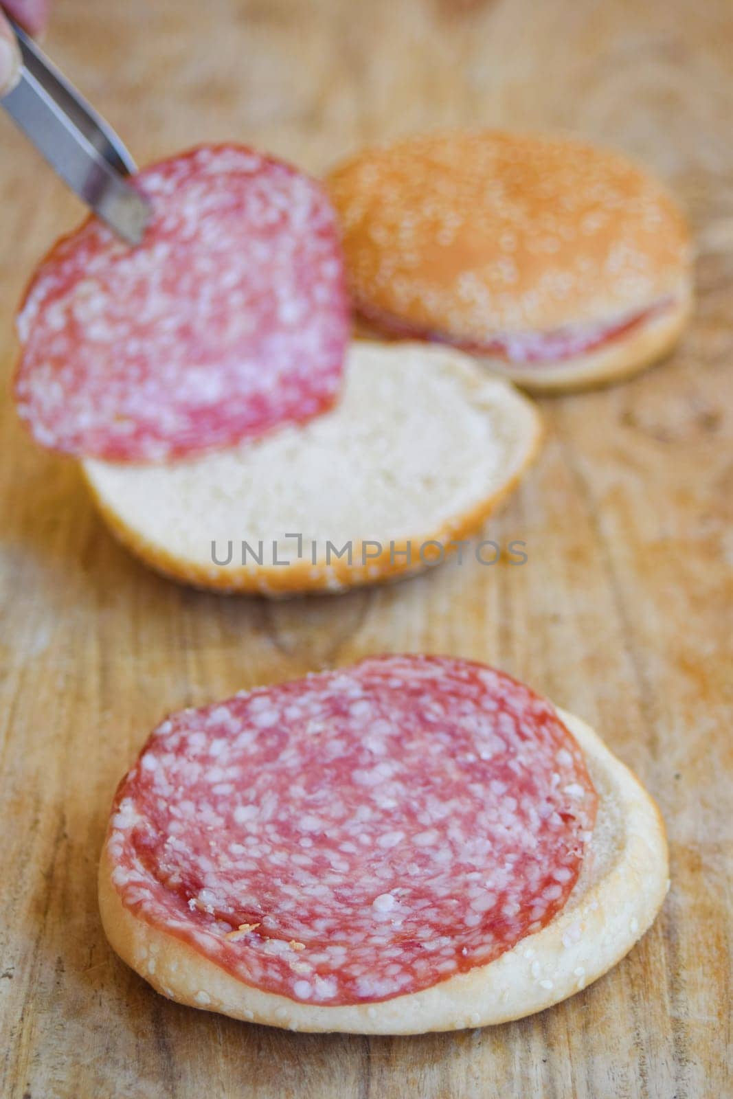 Close-up shot of hands using tongs to place slices of salami on a half of a bread roll, with another assembled salami sandwich in the background on a wooden cutting board