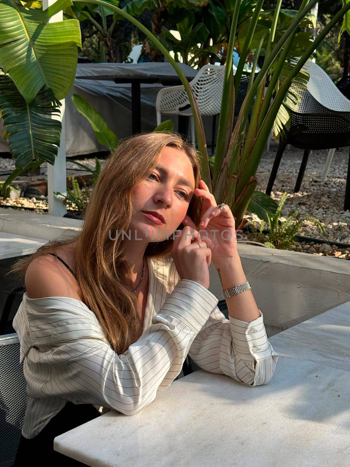 A woman with long hair sits at a marble table in a lush garden, looking contemplative in the warm sunlight of a beautiful afternoon.