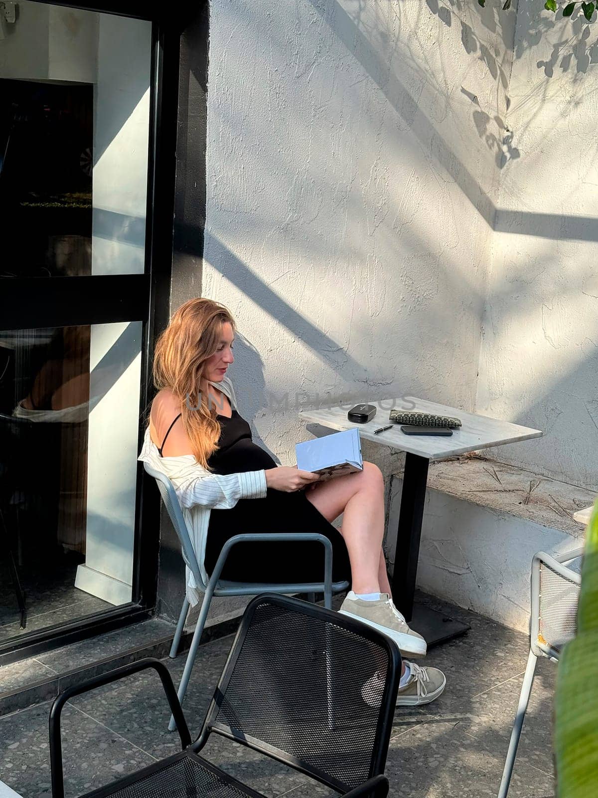 A woman enjoys reading a book while seated in a stylish outdoor cafe during a bright day, showcasing relaxation and leisure.