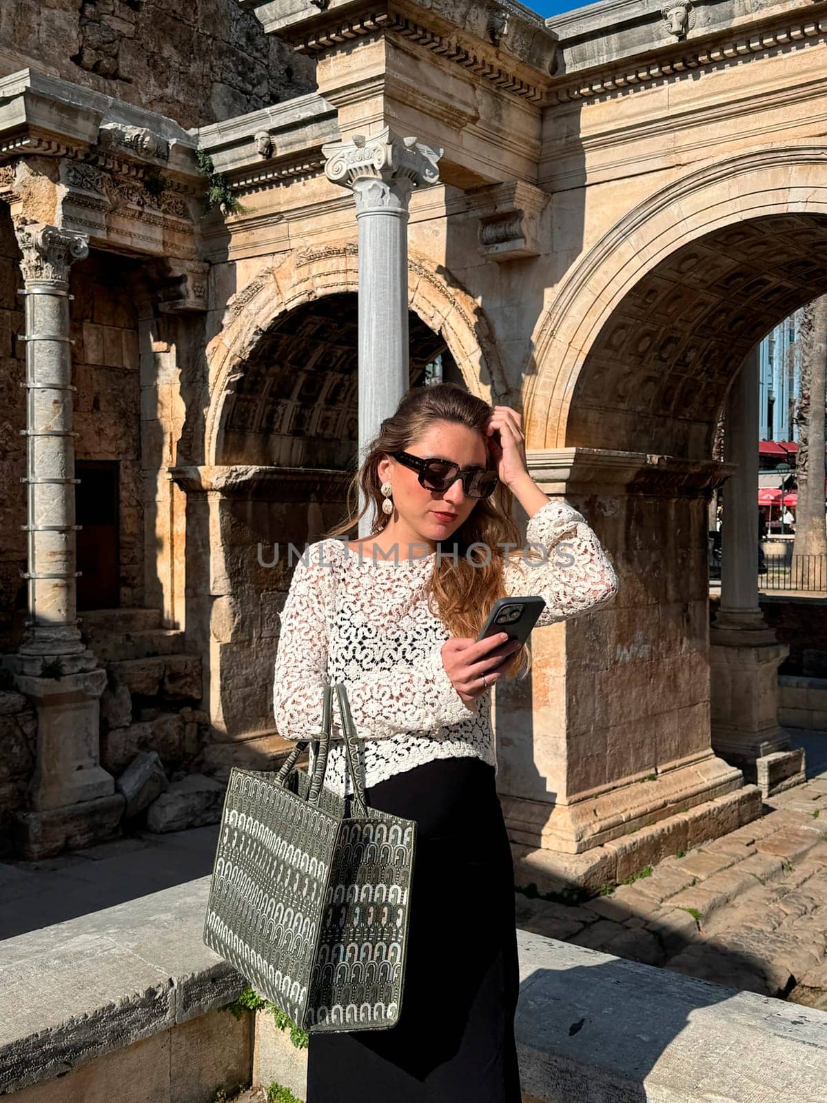A young woman stands by historic stone ruins, checking her smartphone while wearing casual, stylish attire.