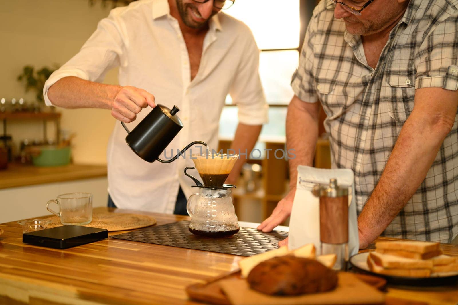Mature man and adult son talk and bond during the process of brewing coffee in kitchen.