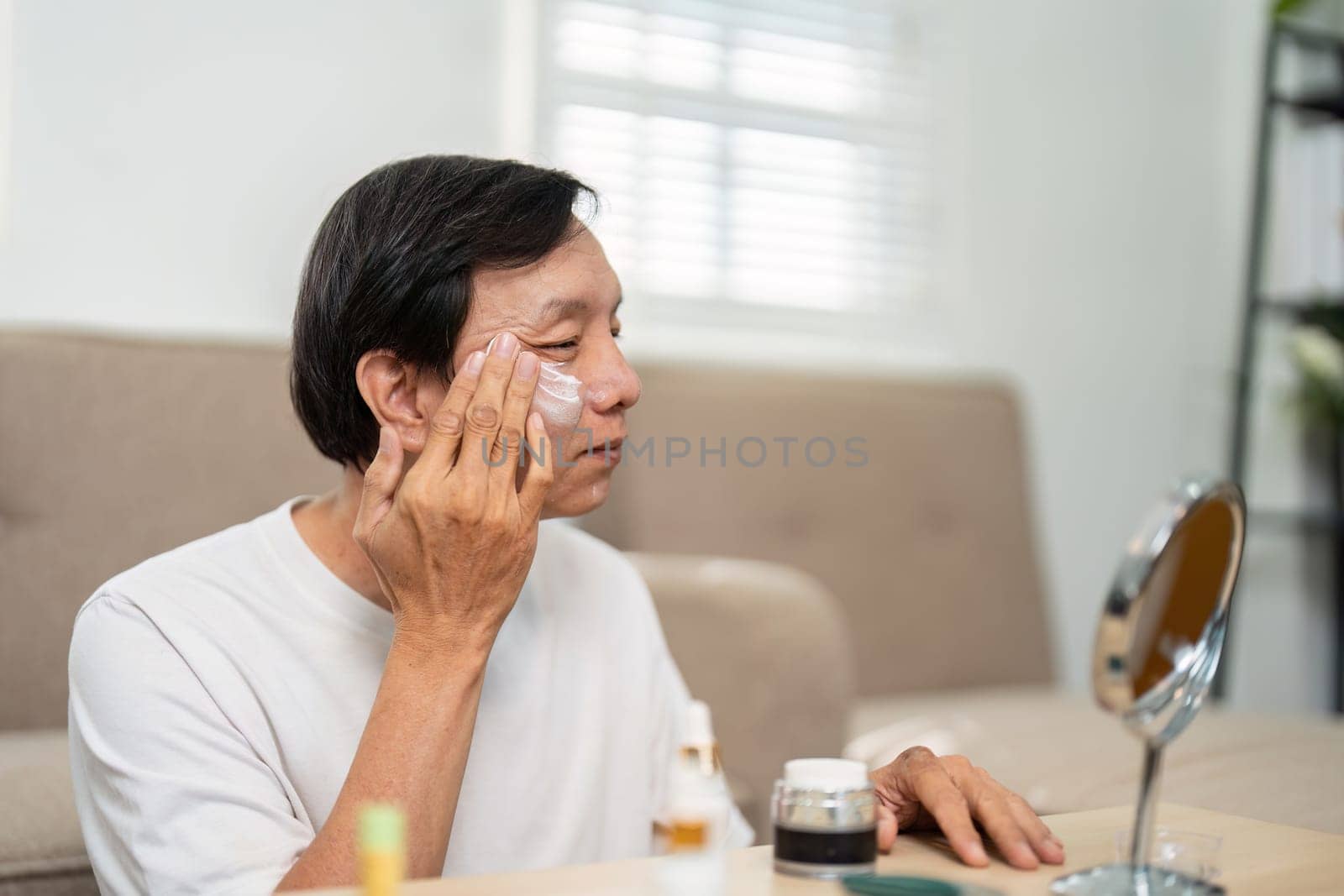 A senior man takes time for his skincare, applying products thoughtfully in front of a mirror, emphasizing the importance of self-care for aging skin.
