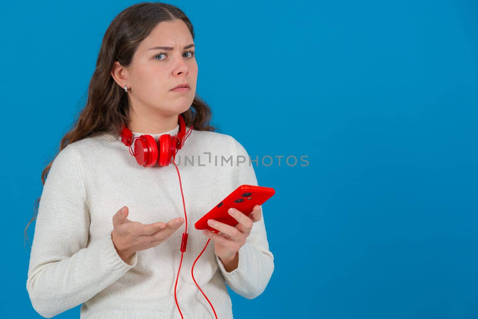 Young woman looking confused and frustrated while holding a red smartphone. Wearing red headphones around her neck, she reacts to a problem or error against a bright blue background. Copy space