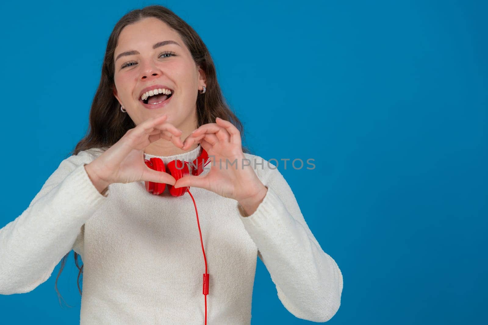 Young woman in white sweater and red headphones smiling while making a heart shape with her hands against a blue background, representing kindness, connection, love, and positive emotions. Copy space