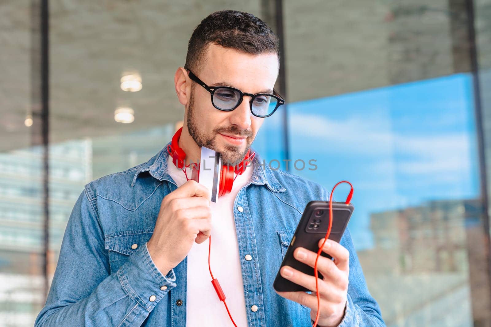 Happy man celebrating a successful online purchase while holding a credit card and smartphone. He smiles with excitement, wearing headphones and sunglasses in a modern urban environment.