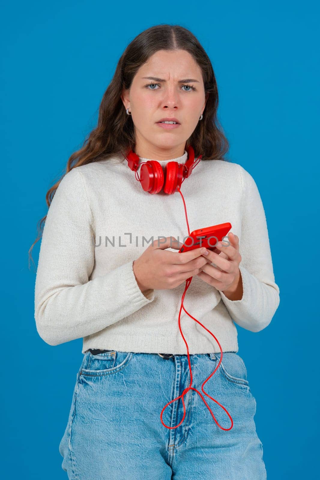 Young woman looking confused and frustrated while holding a red smartphone. Wearing red headphones around her neck, she reacts to a problem or error against a bright blue background. Vertical
