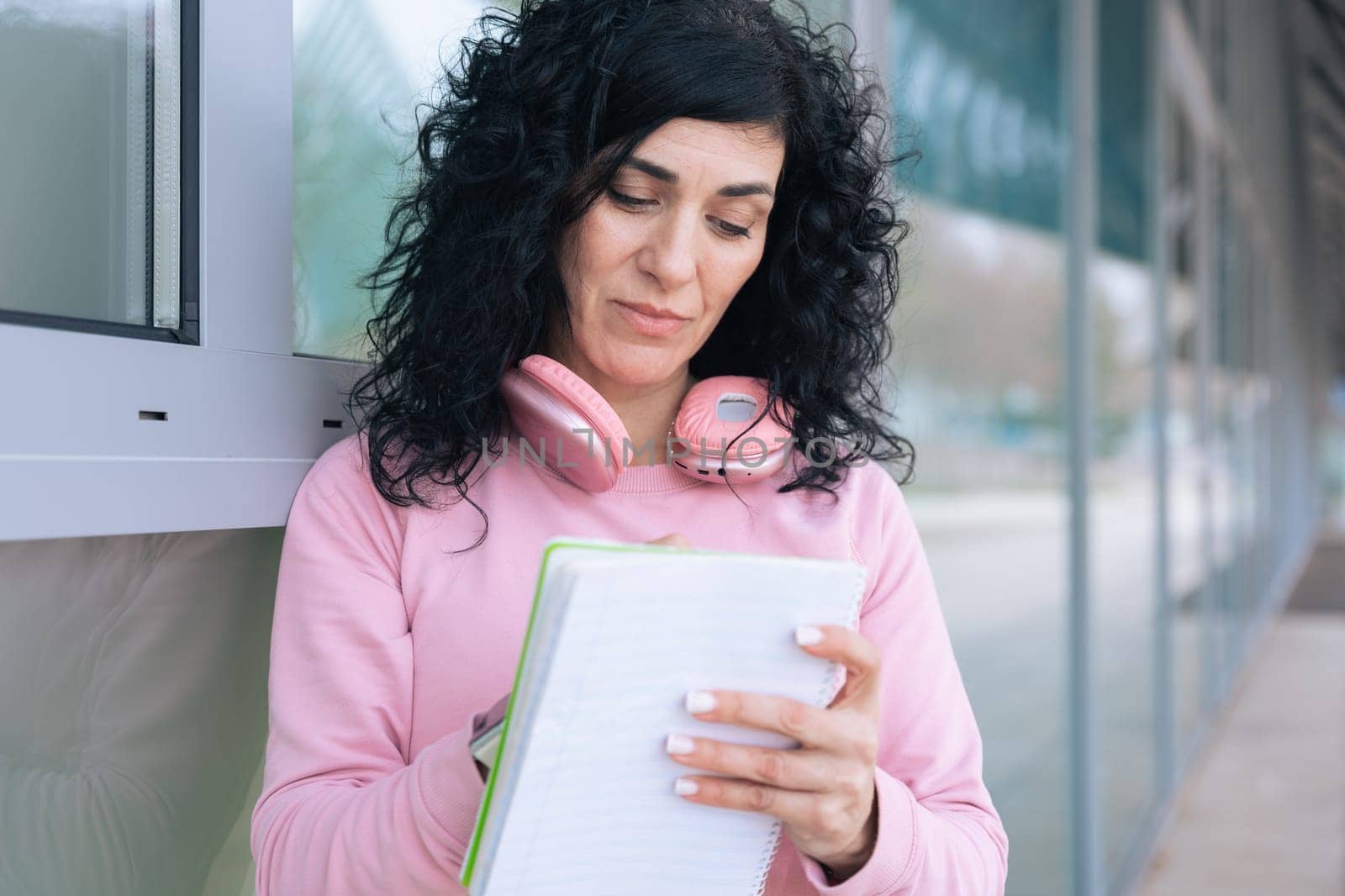 Caucasian sporty woman in a pink hoodie writing in a notebook outdoors. Wearing headphones around her neck, she appears focused and relaxed after a workout in an urban setting.