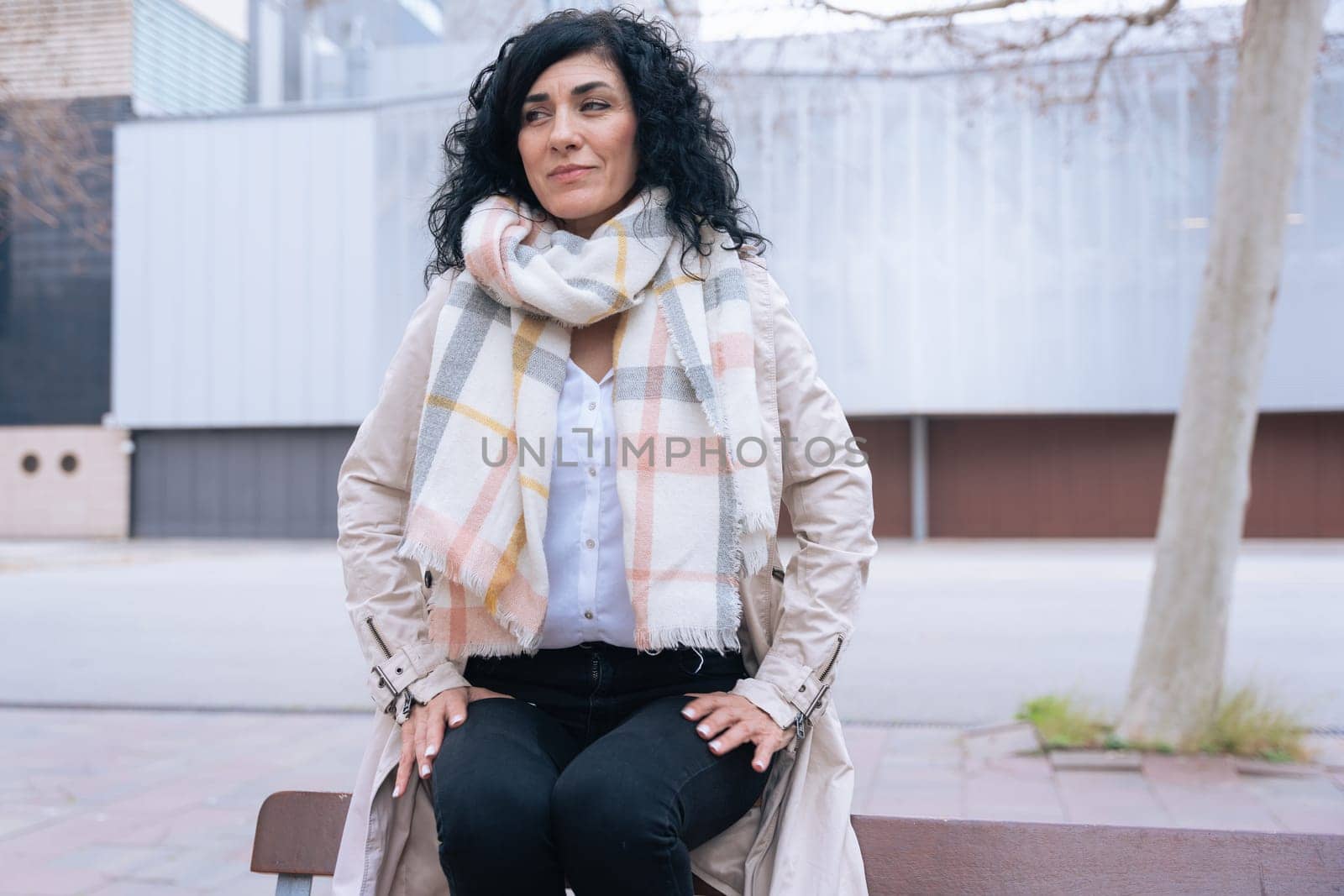 Horizontal portrait of a woman sitting on a bench outdoors. She wears a beige coat and scarf, looking calm and confident. Urban background with winter atmosphere and seasonal fashion concept.