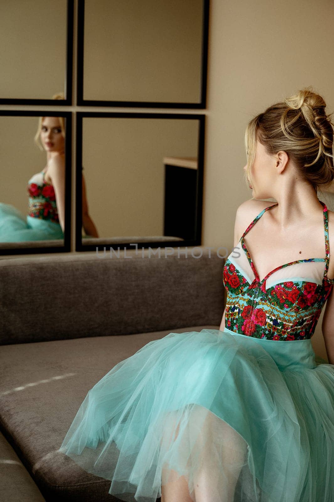 Stylish blonde in bright turquoise dress sits in minimalist interior. Girl has high hair. On the corset bright red embroidery of flowers. She radiates spring energy and joy, looks in the mirror