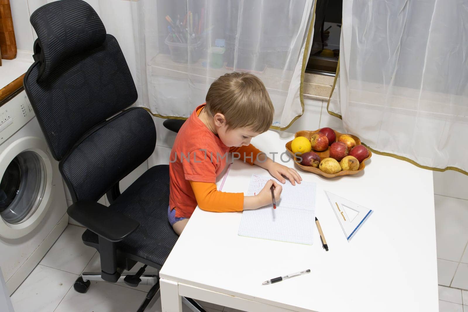 Young boy practicing drawing and writing on notebook, sitting on an ergonomic chair next to a bowl of fresh fruit