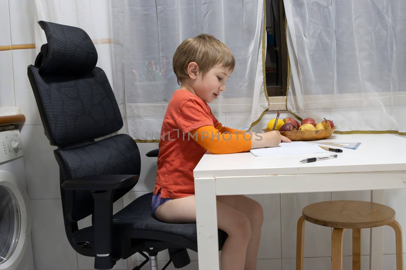 Young boy doing homework, sitting on a big office chair and drawing on a notebook placed on the kitchen table