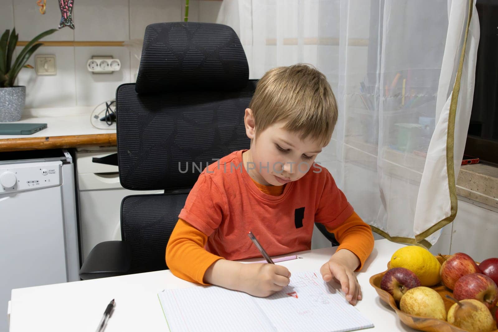 Preschool student writing on notebook sitting at kitchen table, diligently completing homework assignment