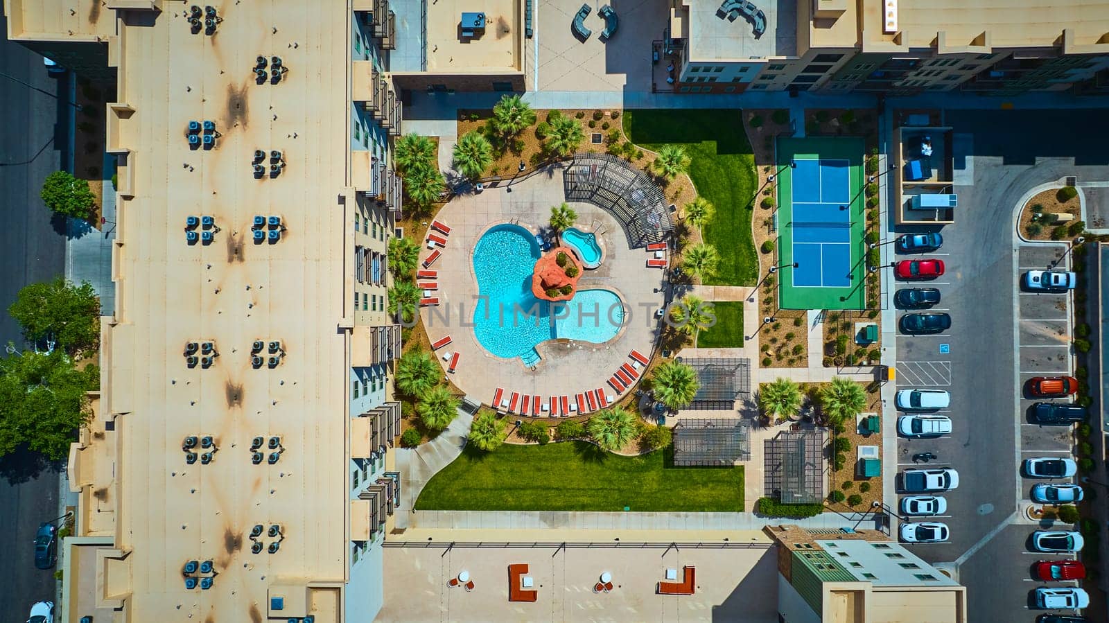Aerial view of a modern St George Utah apartment complex featuring a resort style swimming pool, lush palm landscaping, and a vibrant blue tennis court under bright midday sun for upscale urban living