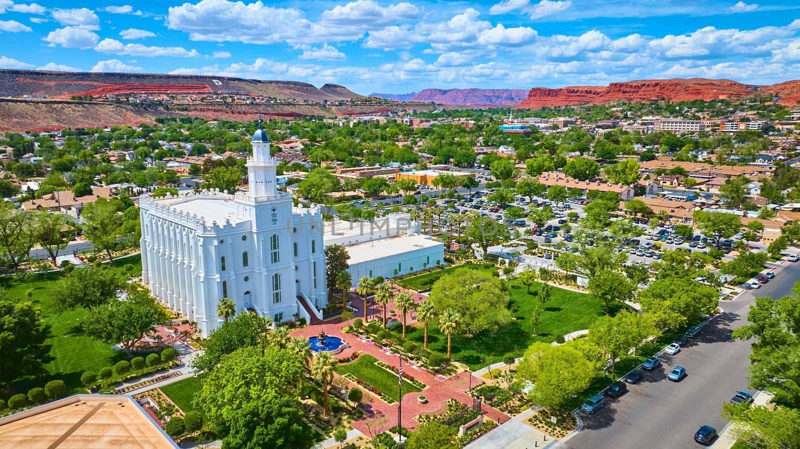 Aerial view of the St George Utah Temple with its striking white architecture surrounded by lush gardens, palm trees, and cityscape set against dramatic red rock cliffs in sunny St George USA