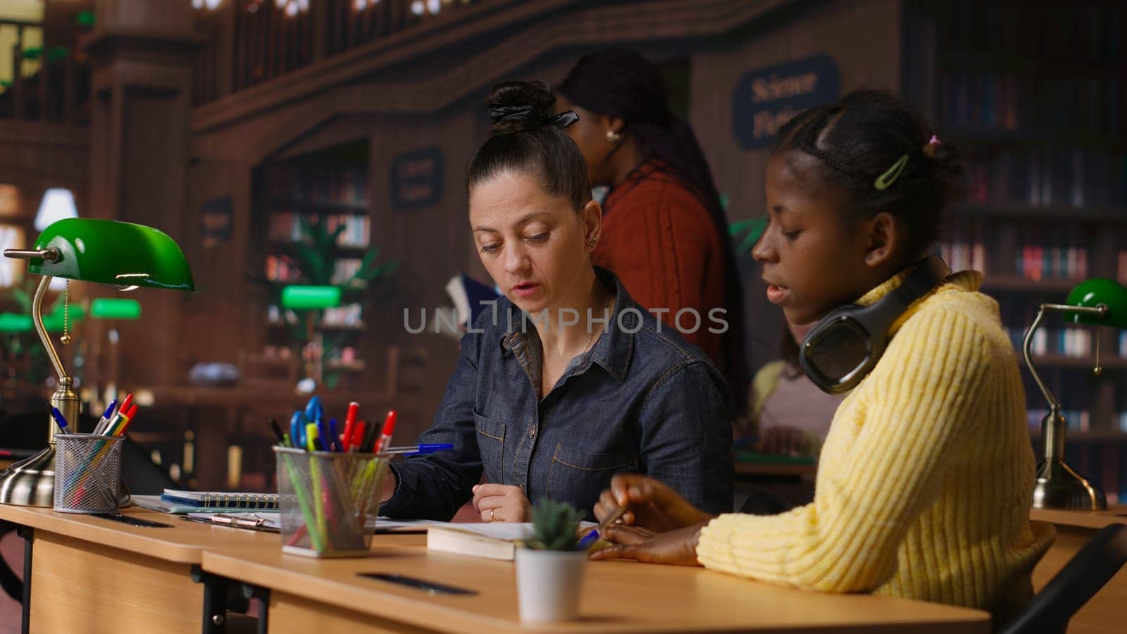 Caucasian tutor guiding elementary school girl at a library desk, providing private lessons to aid in their academic development with work books and notes. Teacher discussing with scholar. Camera B.