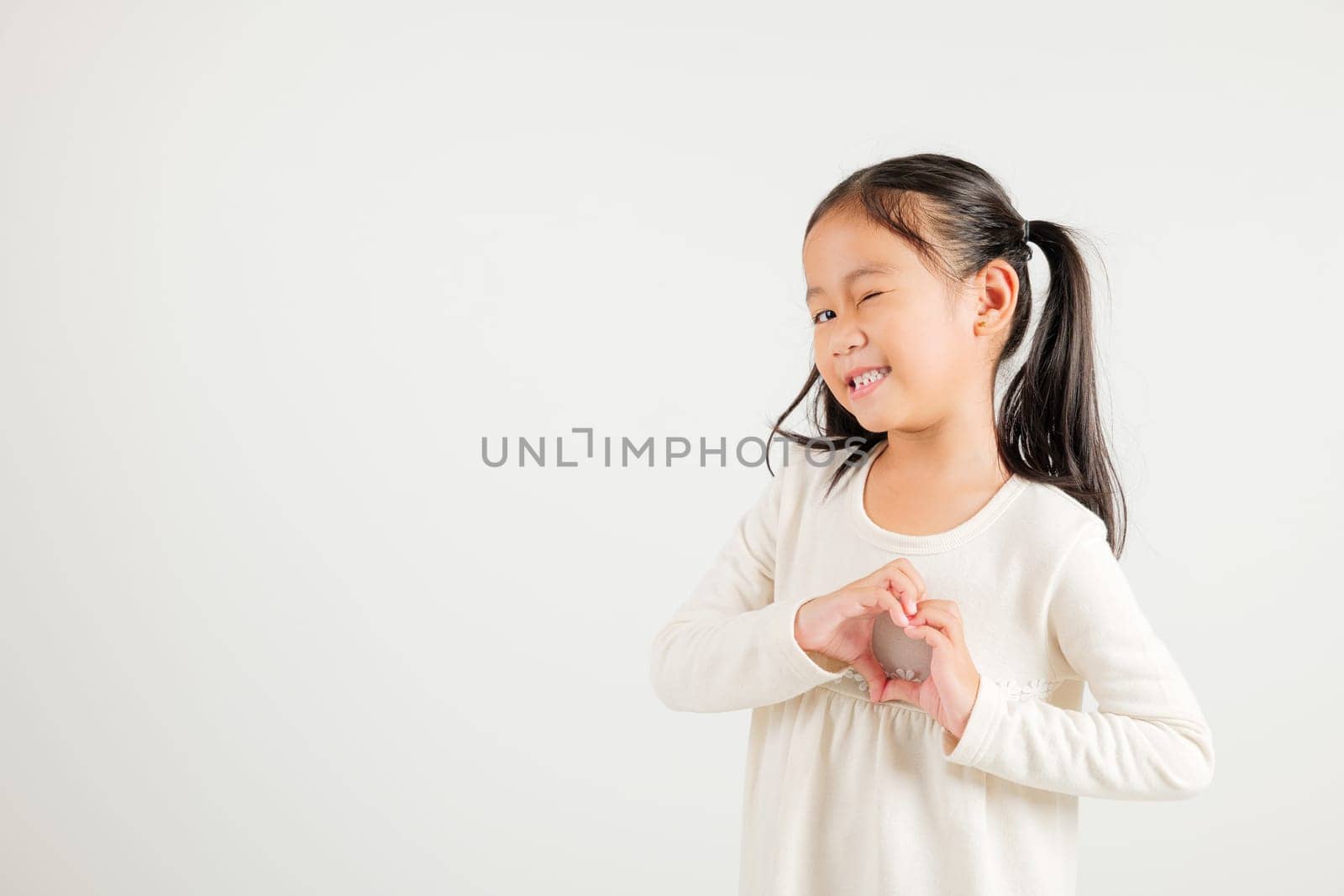 A smiling Asian young kid girl shows confidence, creating a heart shape with fingers and hands on white background. Portrait of kindergarten child sending love for Valentine's Day