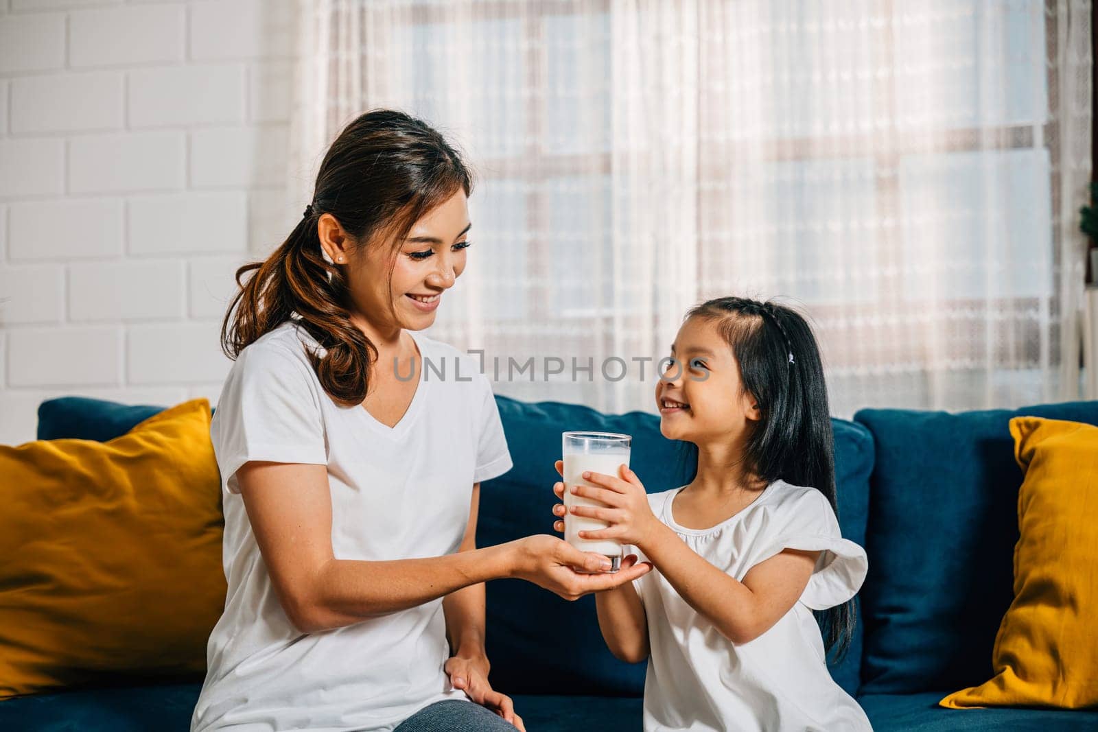 In their modern home an Asian mother lovingly offers her daughter a glass of milk on the sofa. This image reflects their carefree happiness togetherness and calcium-rich diet.