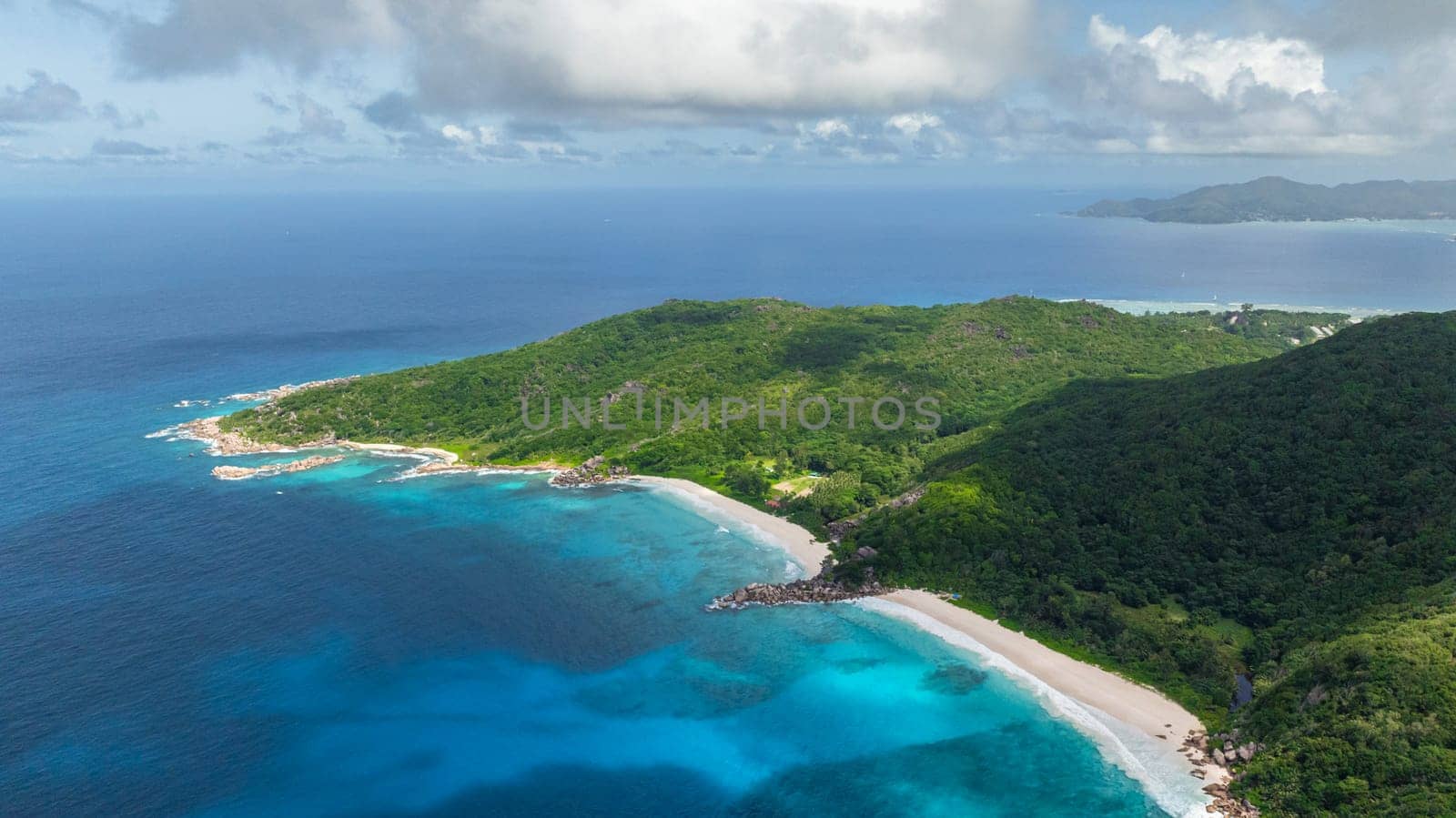 Turquoise ocean waves hitting a rocky shoreline bordered by green forested hills. La Digue, Seychelles.