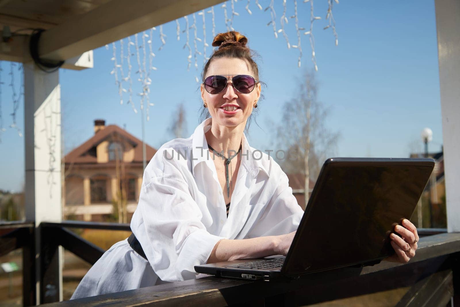 A woman sits at an outdoor table, using her laptop while enjoying the sun, wearing sunglasses and a relaxed outfit in a vibrant setting.
