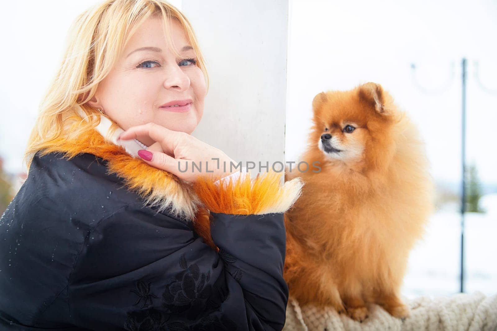 A blonde woman stands next to her fluffy Pomeranian dog, enjoying a serene winter walk in the snow.
