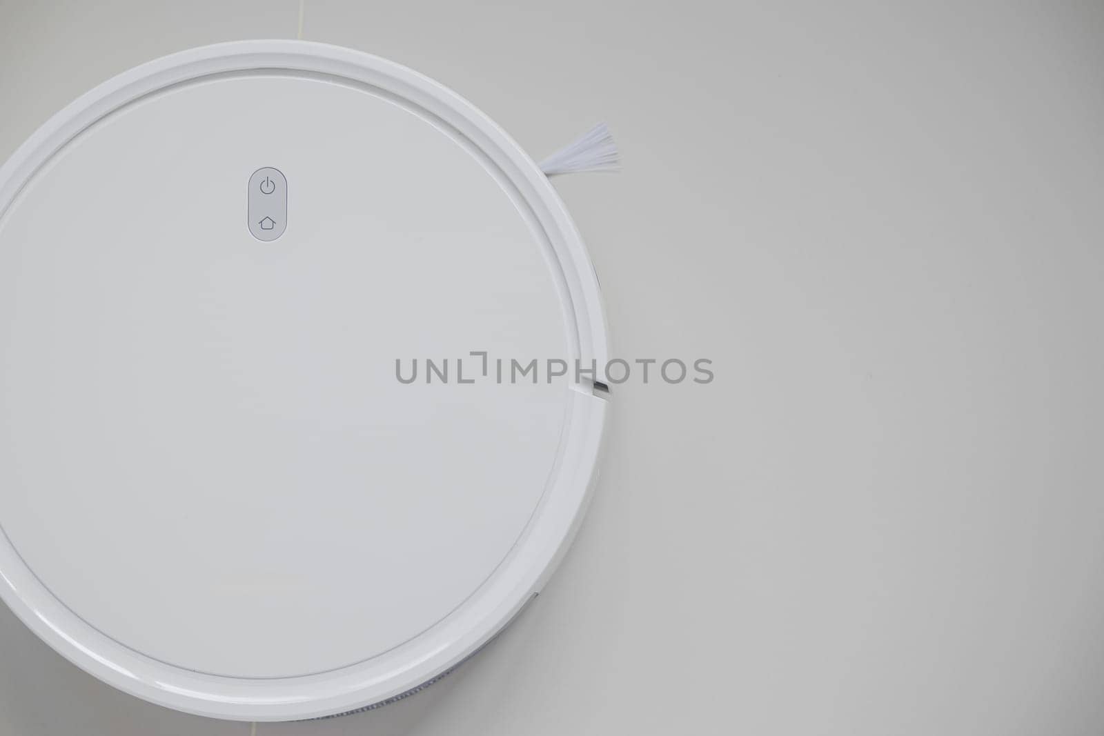 White automatic floor cleaner with side brush on tile flooring. Top view of robot vacuum cleaner removing dust, showcasing modern smart home cleaning technology for spotless and efficient maintenance