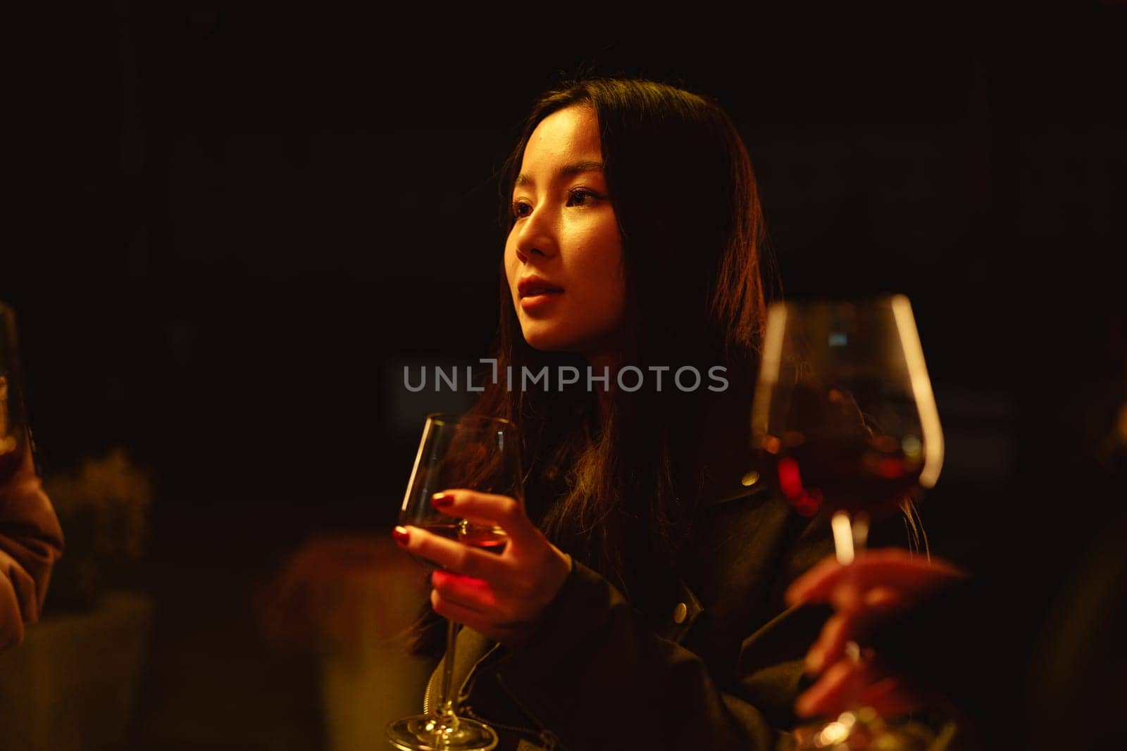 In a cozy venue with soft, warm lighting, a young woman holds a glass of wine and engages in conversation with others nearby, creating a relaxed atmosphere.