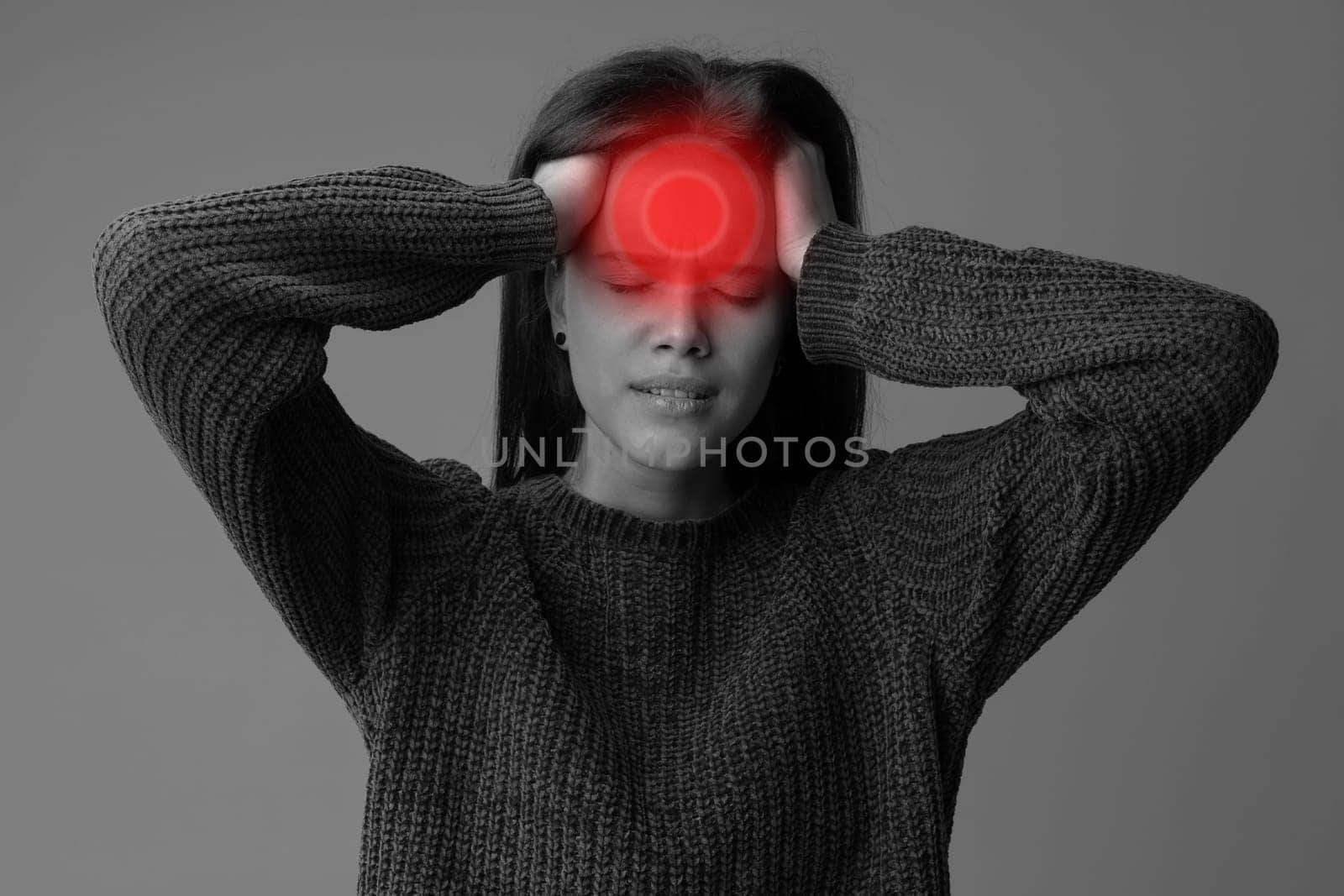 A young woman in a knitted sweater holds her head with both hands, expressing discomfort. She appears to be suffering from a headache, surrounded by a neutral background.