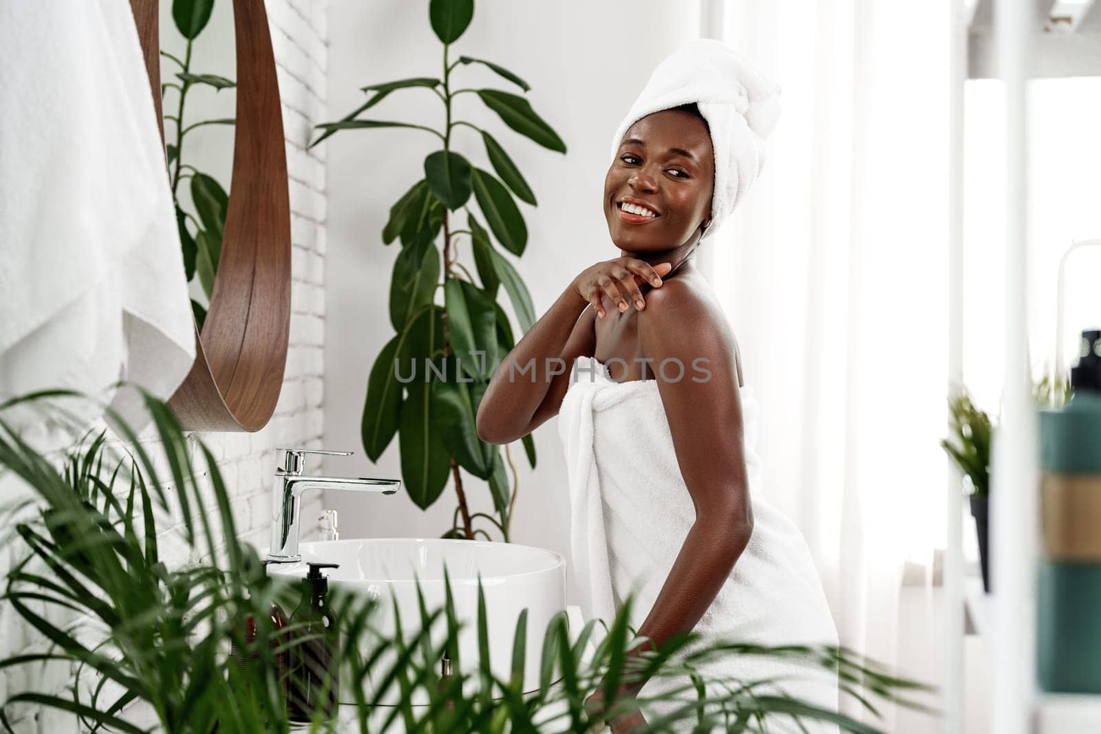 A joyful woman with a towel around her body smiles while applying lotion in a stylish bathroom filled with plants and bright sunshine. The atmosphere exudes tranquility and wellness.
