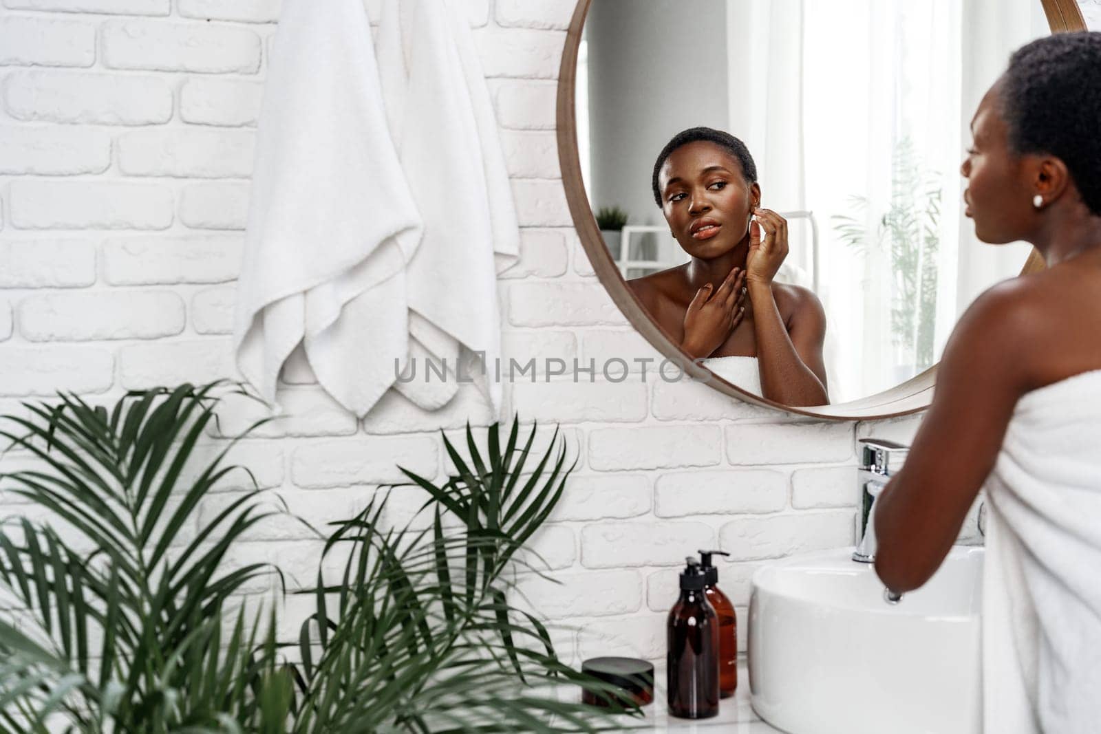 Young woman with short hair applies skincare products while looking in a round mirror. The bright bathroom features white brick walls and lush indoor plants, creating a refreshing atmosphere.