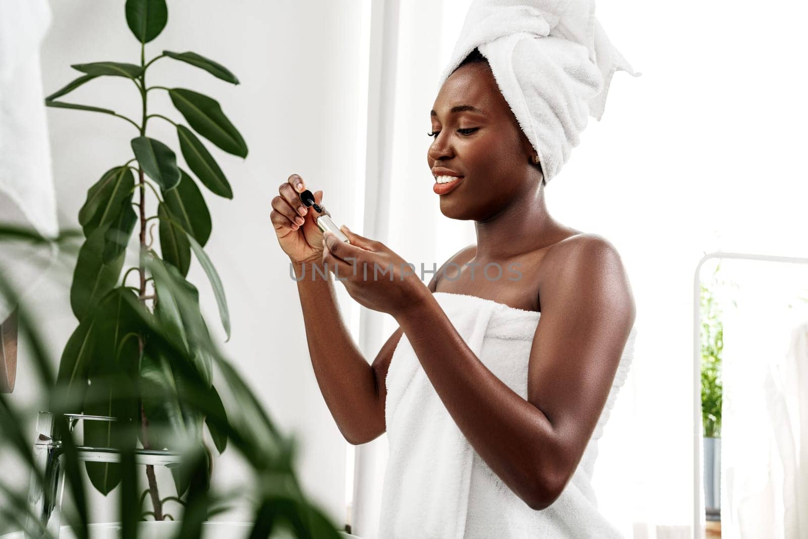 A woman wrapped in a towel smiles as she applies a skincare serum in a well-lit bathroom. Green plants add a natural touch to her relaxing self-care moment.
