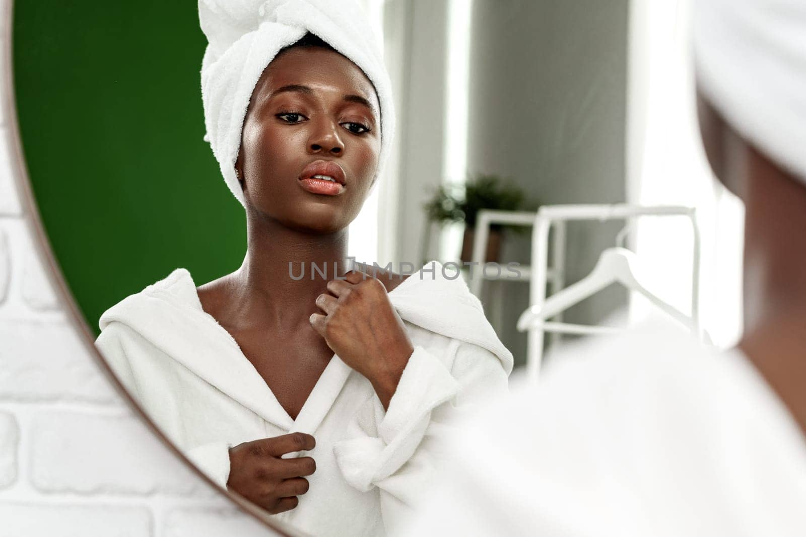 A young woman stands in front of a stylish mirror, wearing a white bathrobe and towel. She adjusts her head towel while gazing thoughtfully, surrounded by a clean, bright bathroom.