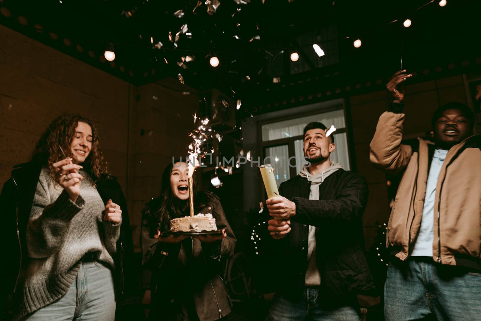 Group of friends gather indoors for a birthday celebration, holding a cake adorned with sparkler candles. They display excitement while enjoying the festive atmosphere, surrounded by decorations.
