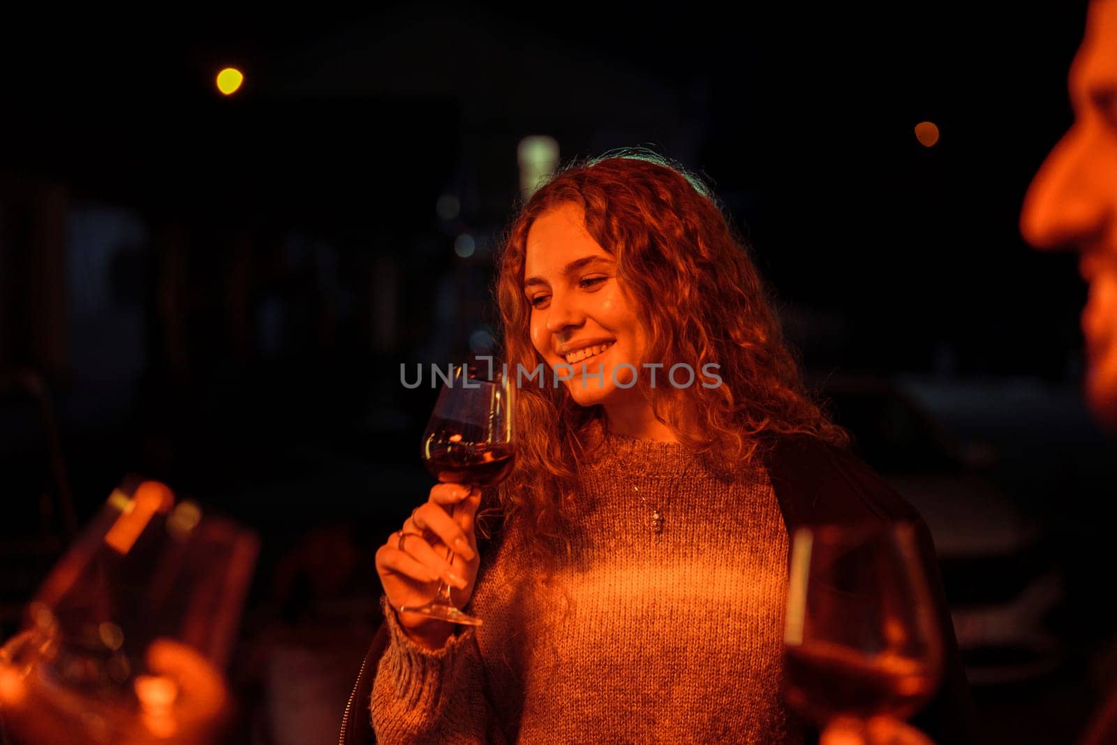 Friends gather on a cool evening, mingling and enjoying glasses of wine. Warm lights cast a cozy glow, enhancing the cheerful atmosphere of their outdoor get-together.