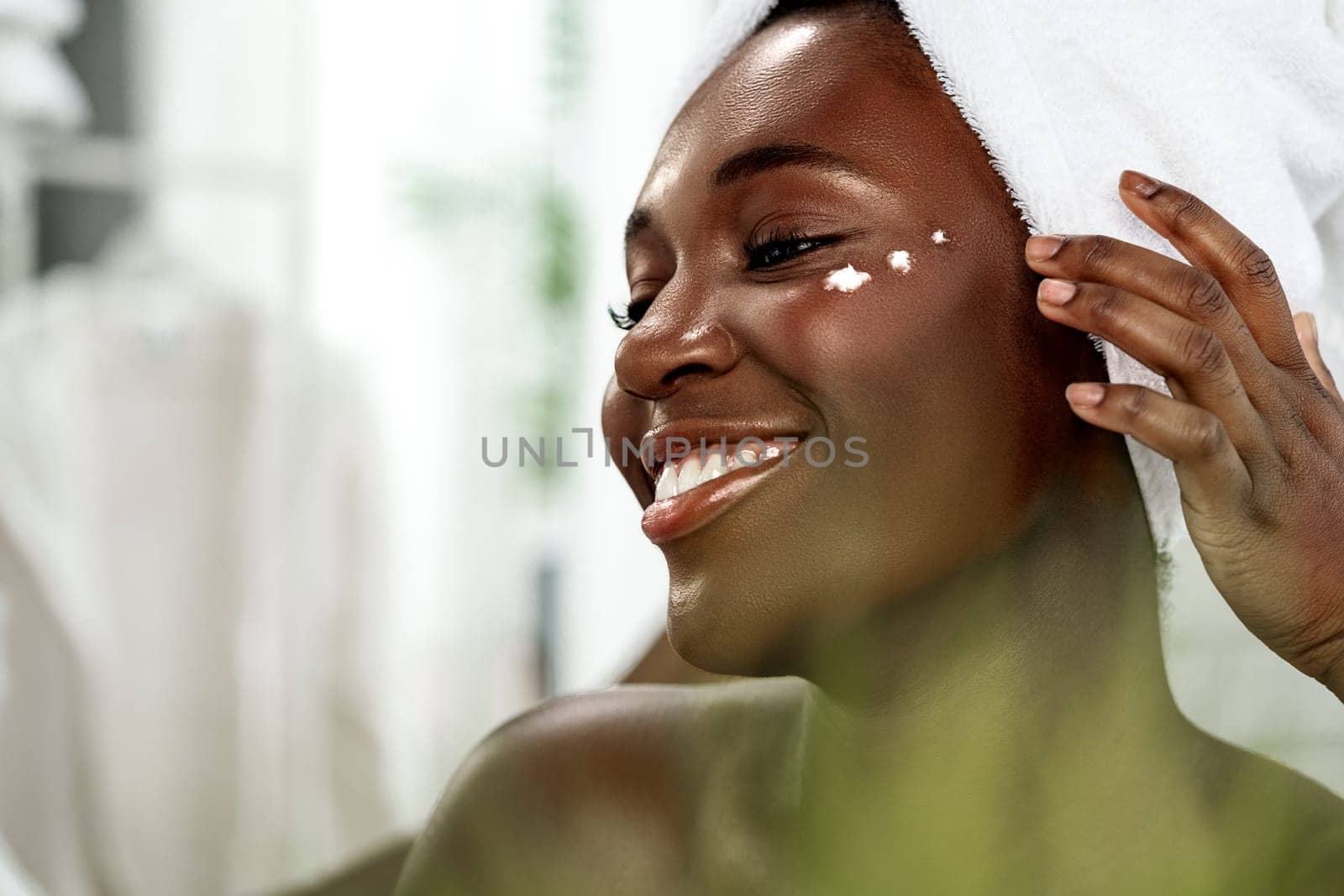 A happy woman with a towel on her head smiles while applying a skincare cream on her cheek in a bright bathroom. Natural light enhances the fresh atmosphere of her morning routine.