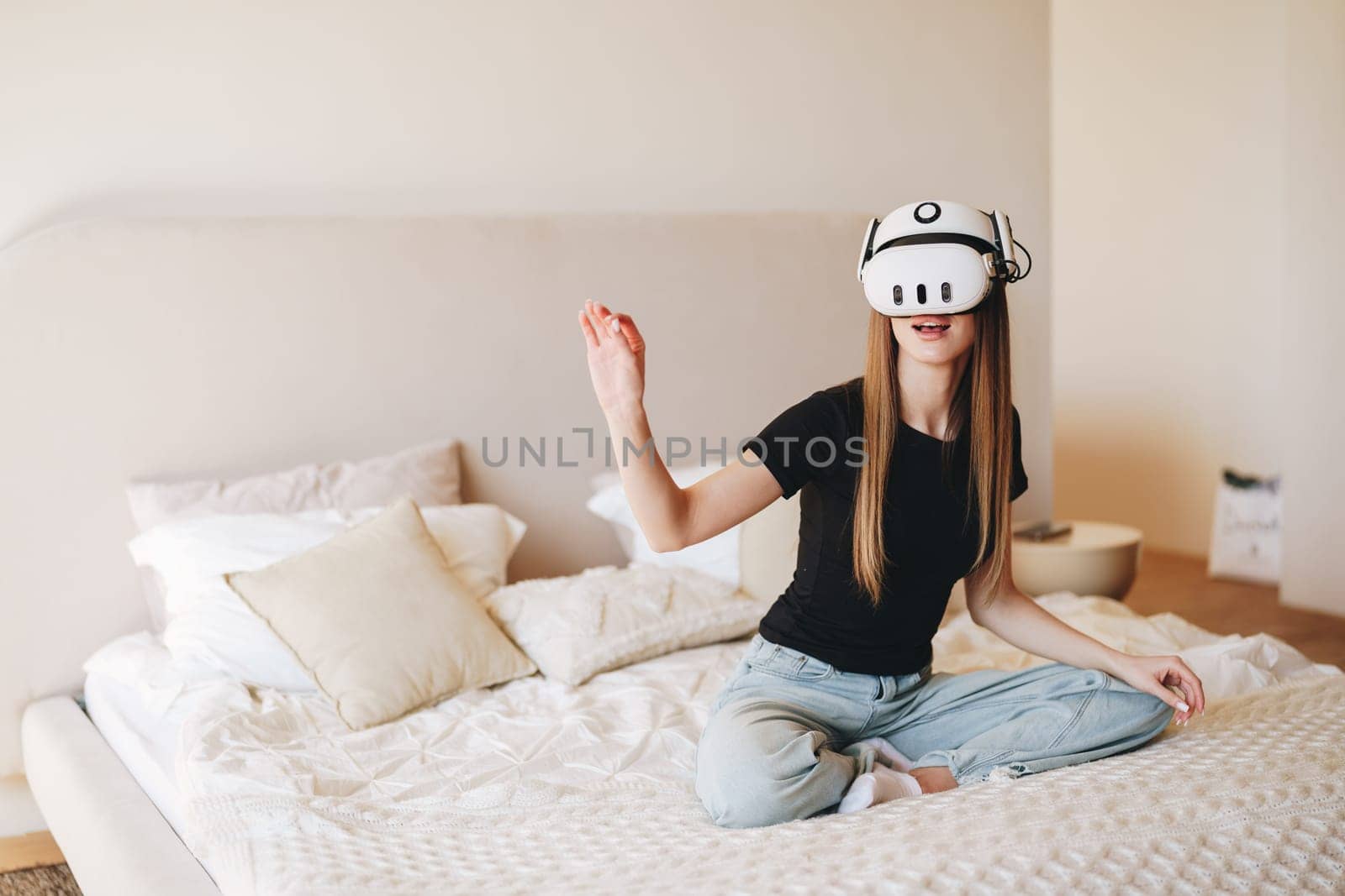 A young woman enjoys a virtual reality experience while seated on a bed adorned with soft bedding in a bright and airy bedroom. Natural light fills the space.