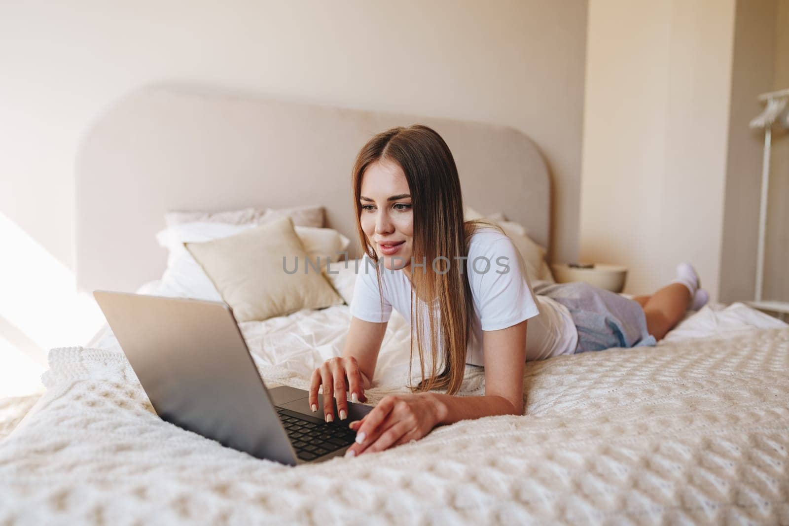 A young woman casually lies on her bed, focused on her laptop. Sunlight filters through the window, illuminating the cozy atmosphere of her serene bedroom.
