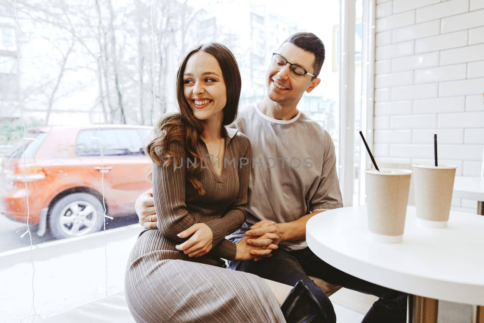 A couple shares a joyful moment in a bright cafe, smiling and looking at each other while seated at a small table. Two cups sit nearby, adding to the warm atmosphere.
