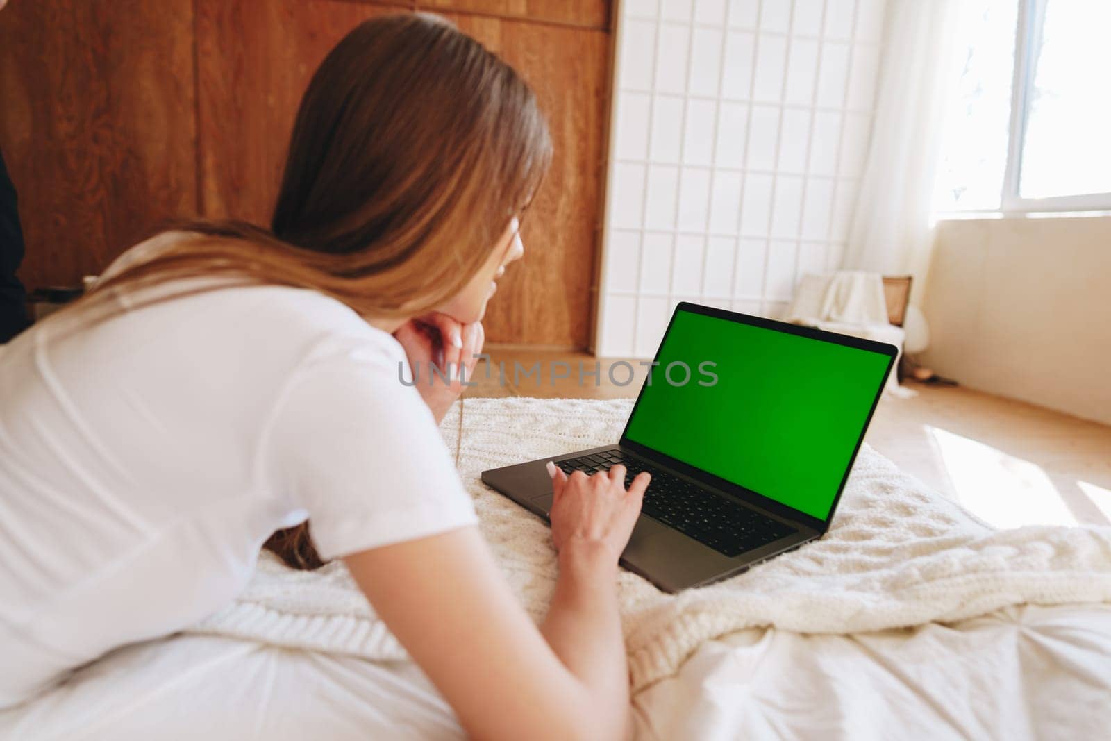 A woman is focused on her laptop in a well-lit room. She lies on a comfortable bed, using her device to complete tasks in a peaceful atmosphere. Sunlight streams through the window.
