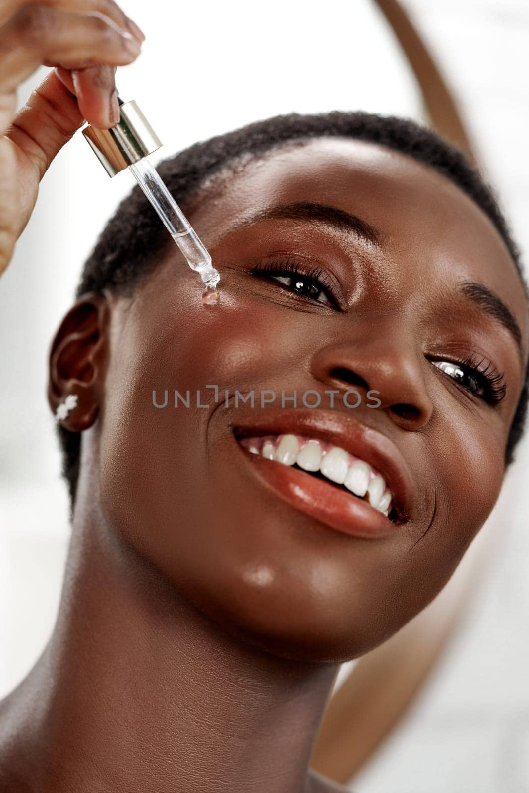 A woman with short hair smiles while applying a serum to her cheek using a dropper. The modern bathroom features natural lighting, enhancing her radiant skin.