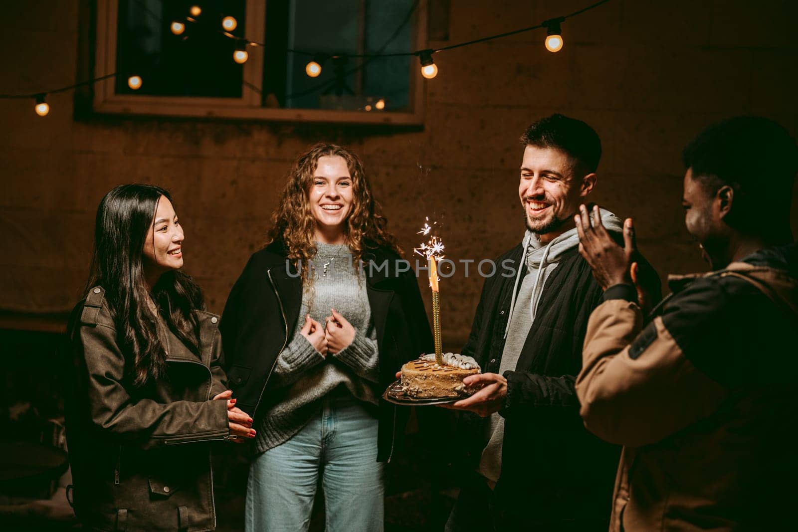 Friends gather outdoors to celebrate a birthday during the evening. The atmosphere is lively and festive, featuring a cake with a sparkler while they enjoy each other's company.