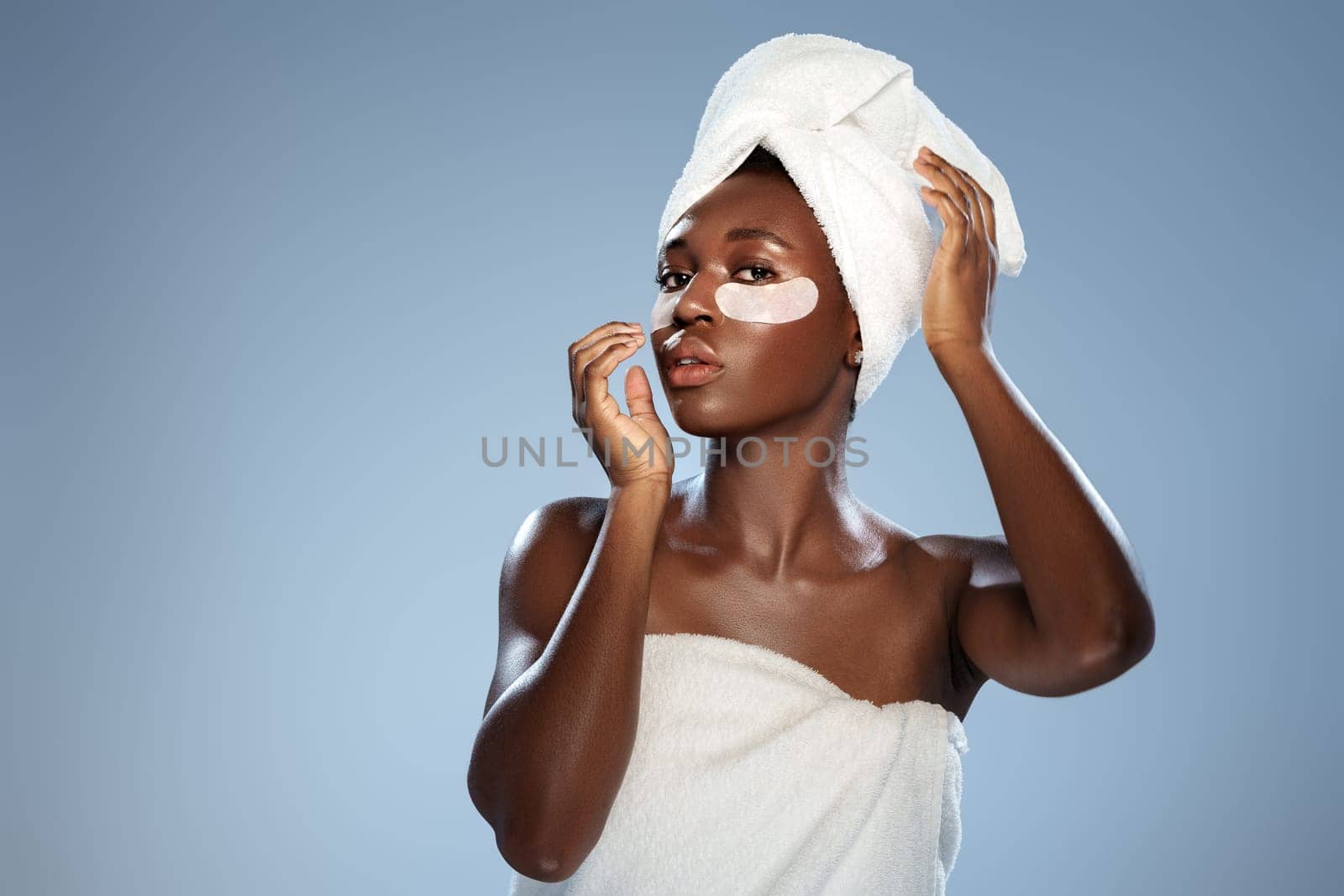 A young woman stands in a serene indoor space, draped in a towel, applying a skincare treatment under her eyes. She exhibits a calm and focused expression, showcasing her self-care routine.