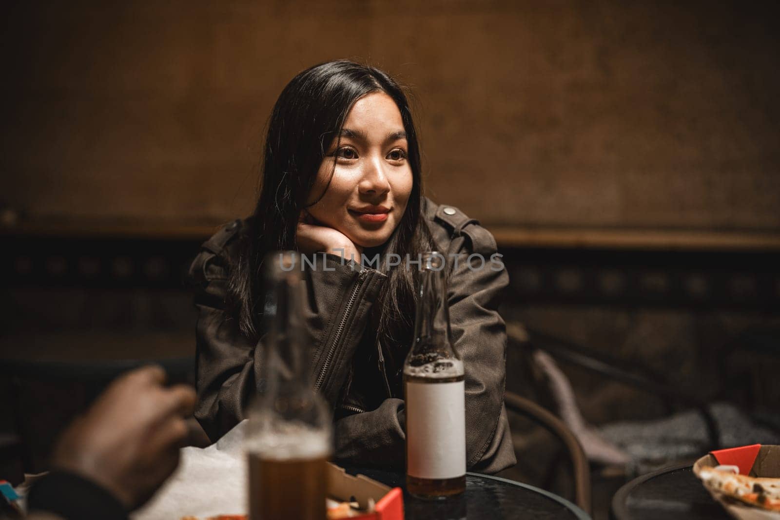 In a cozy atmosphere, a young woman rests her chin on her hand, deep in thought while surrounded by friends and drinks. The soft lighting creates an intimate vibe perfect for conversation.