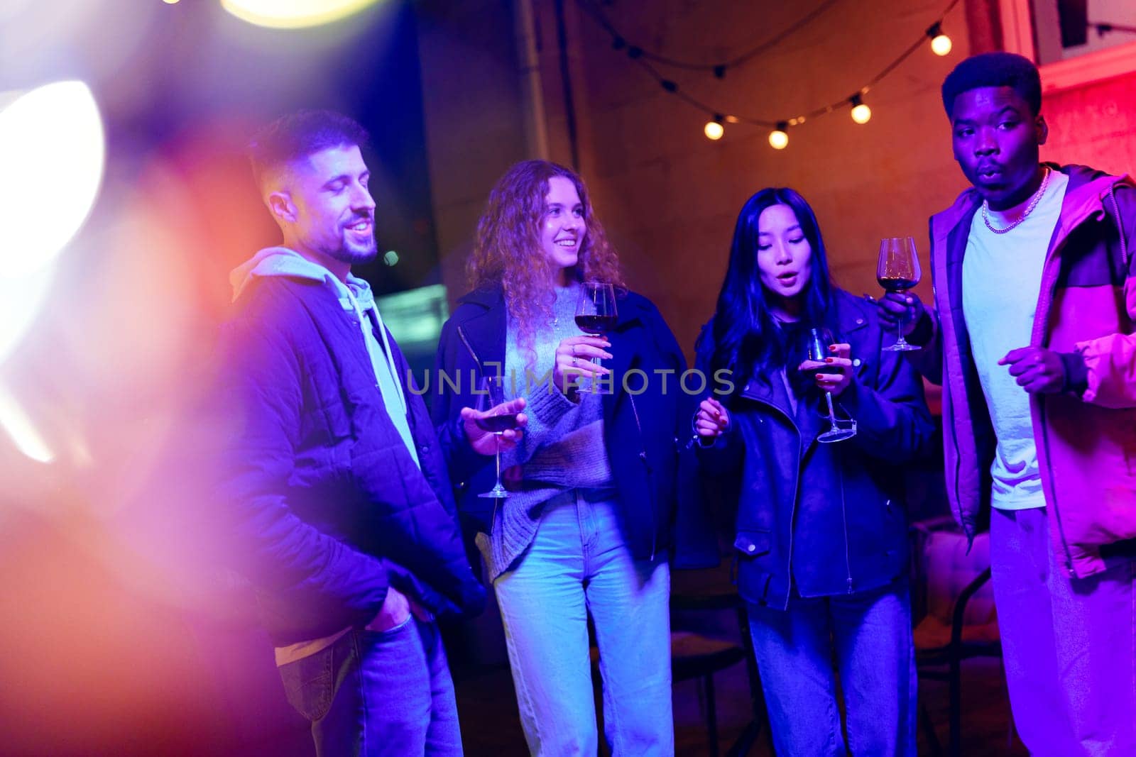 Four friends gather outdoors, holding wine glasses and smiling under vibrant lights. The cozy atmosphere fosters laughter and conversation on a lively night.