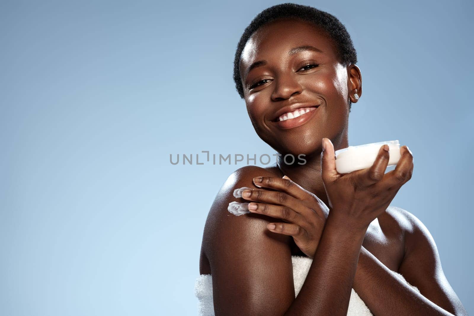 A woman with short hair smiles broadly while applying a skincare product to her shoulder, showcasing her radiant skin in a bright, light blue setting.
