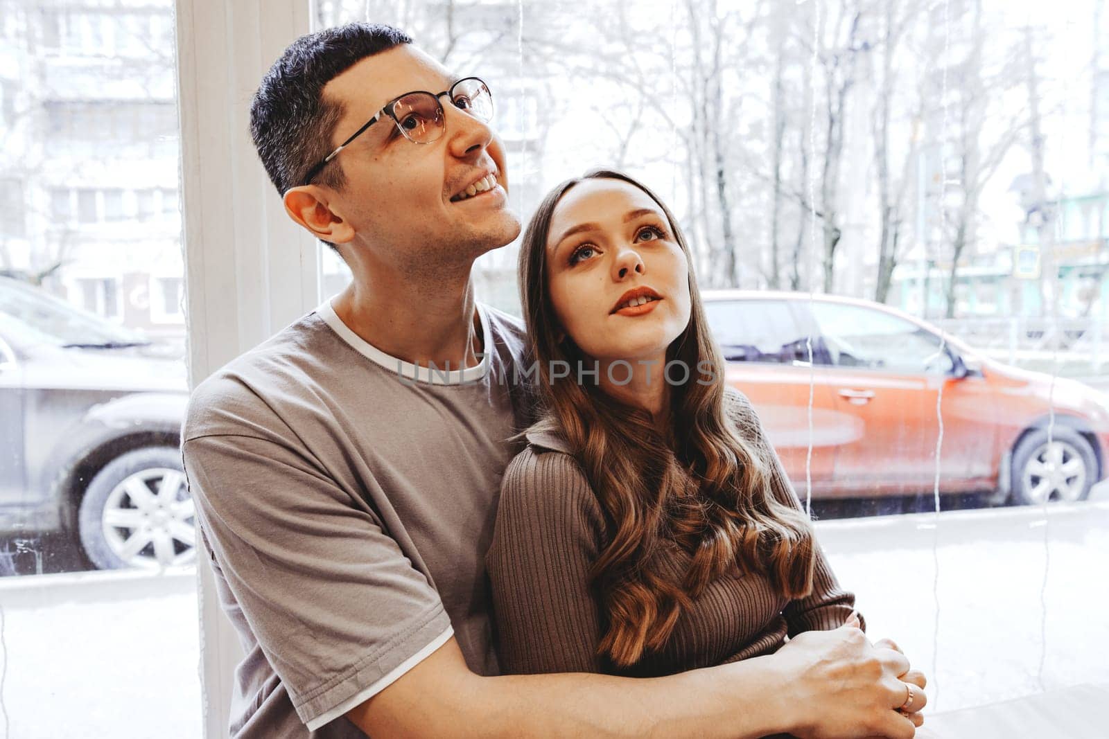 A man and woman sit closely together in a warm cafe, embracing each other with smiles, as rain falls outside, creating a cozy atmosphere perfect for connecting.