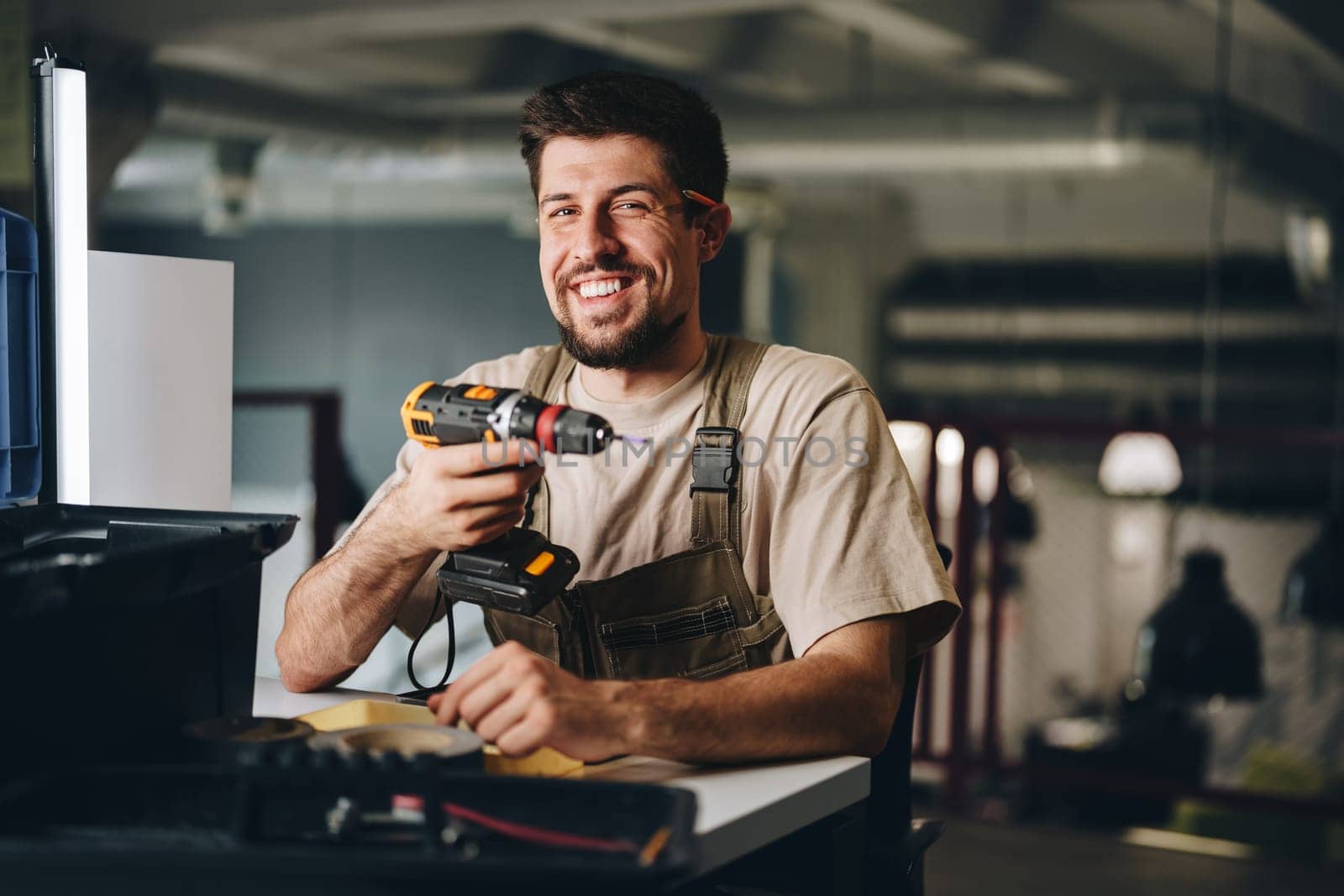 A skilled worker with a friendly smile holds a power drill in a modern workshop. Tools and materials are neatly arranged, showcasing a productive atmosphere.