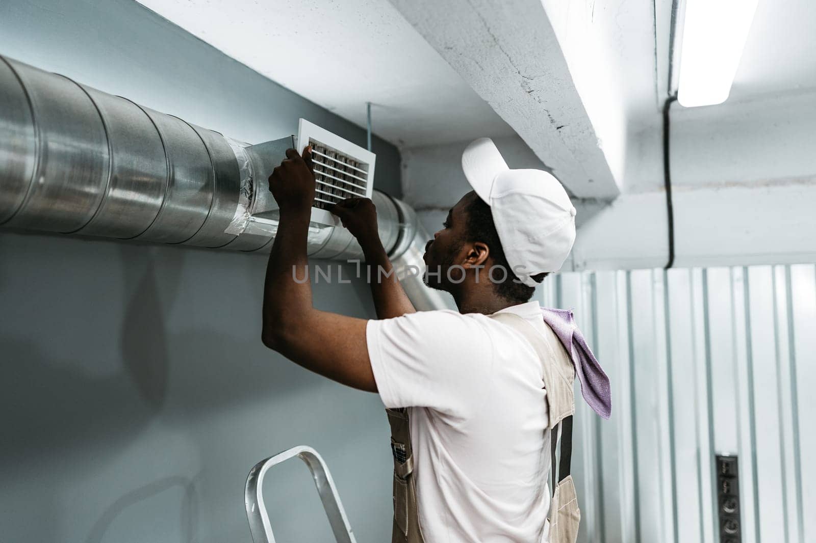 A technician adjusts an air vent while standing on a ladder in a commercial space. The setting features metal ductwork and ambient lighting. Attention to detail is evident in the careful work.