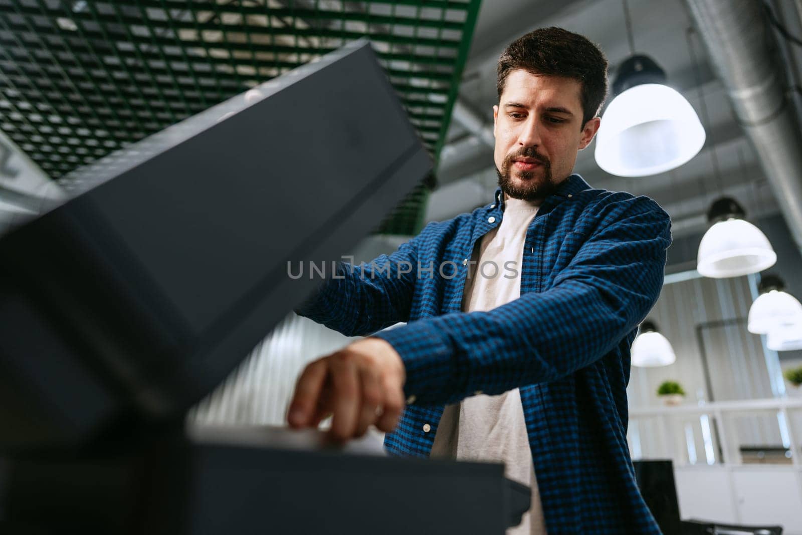 A young man in a blue shirt interacts with a sleek office printer, attentively managing the printing process in a contemporary workspace filled with soft lighting.