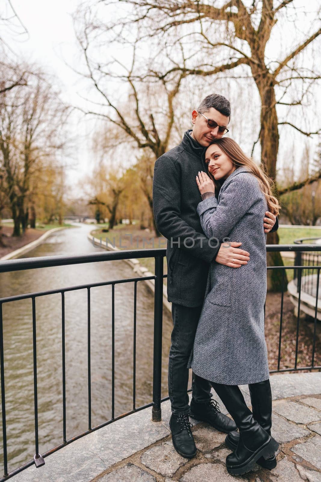 In a tranquil park setting, a couple enjoys an affectionate moment by the river. The trees are bare, and soft light enhances the cozy atmosphere of their embrace.